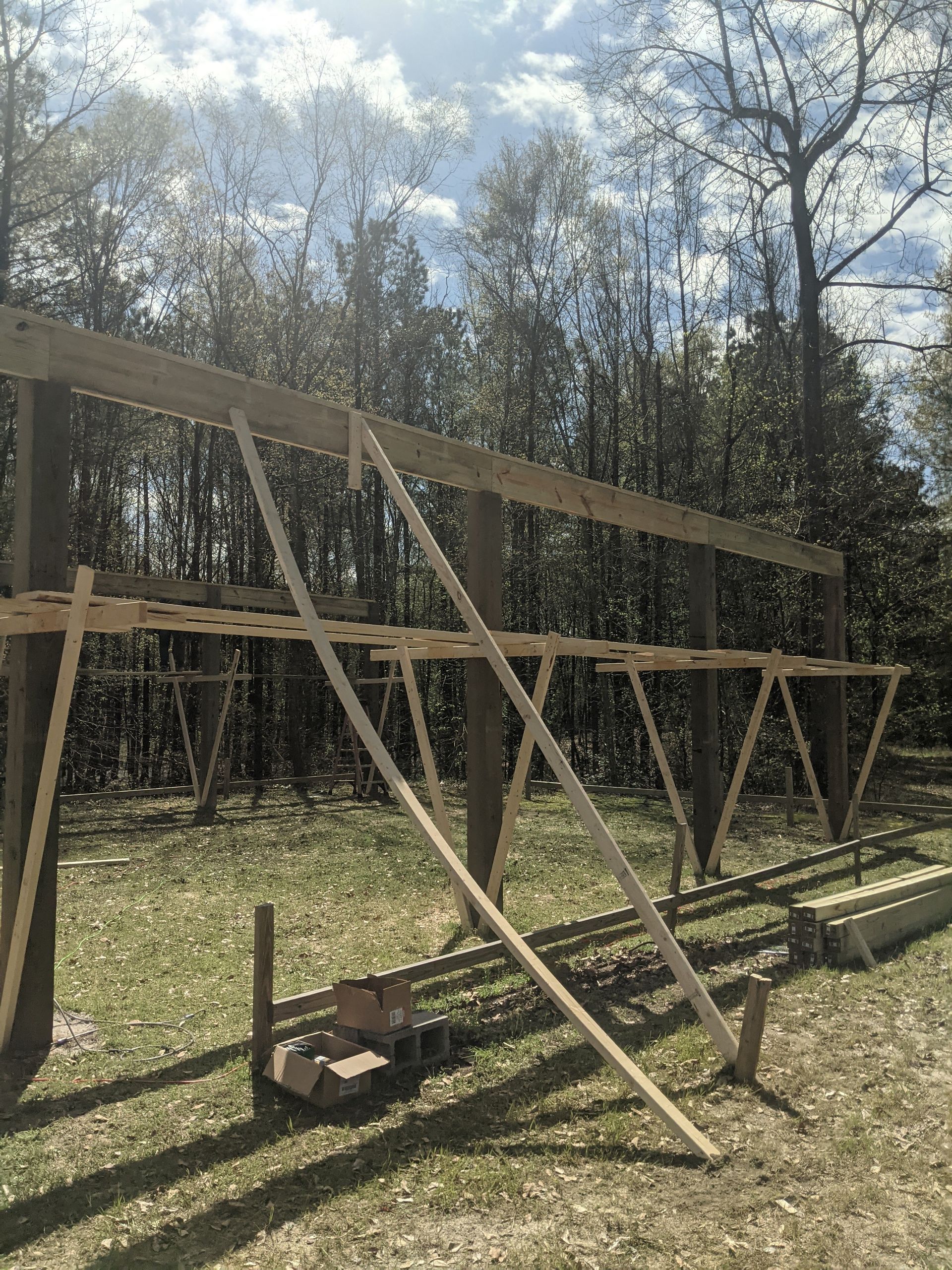 A wooden frame for a building project stands in a wooded area, supported by diagonal braces against vertical posts.