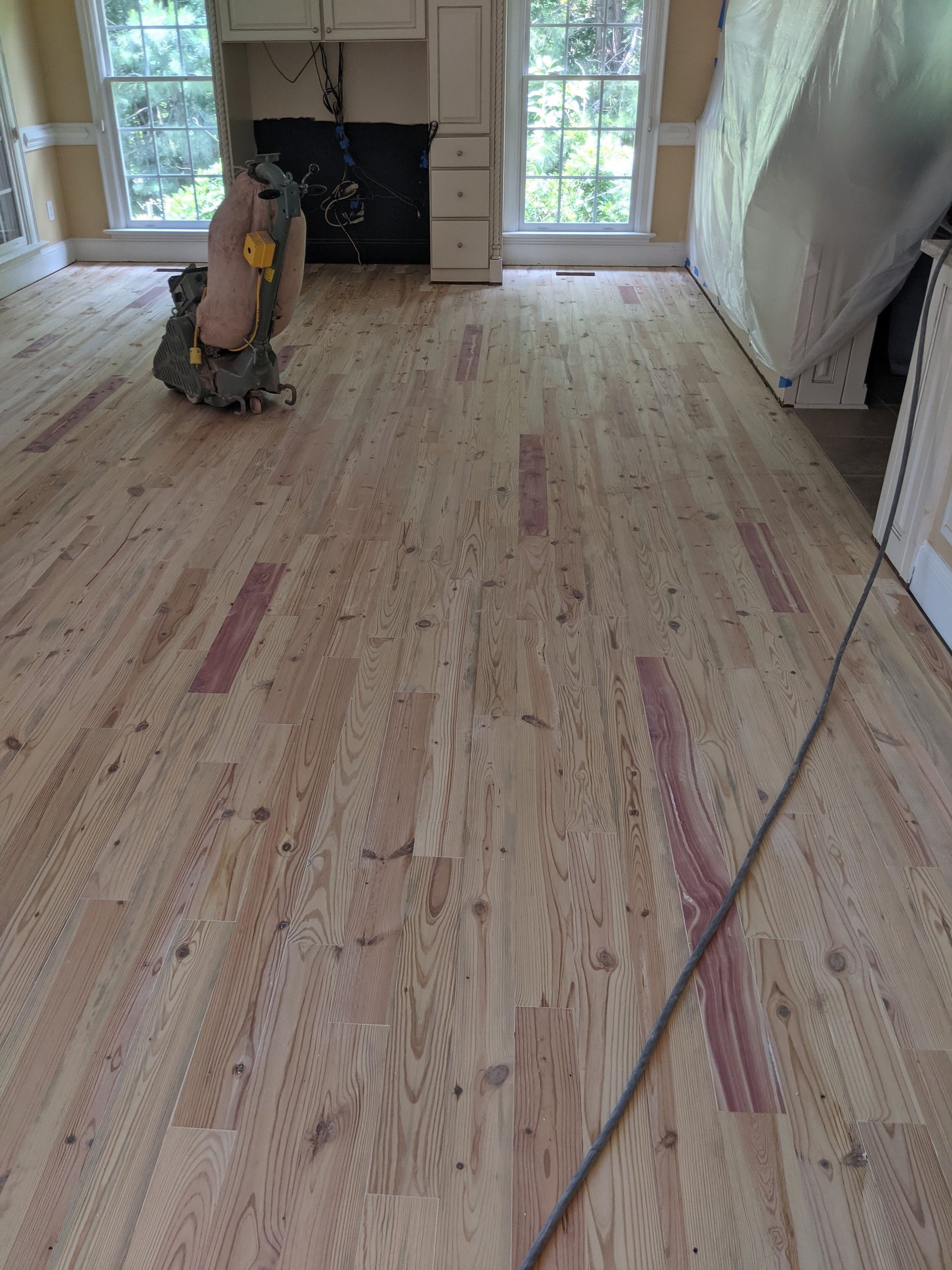 A room with unfinished, sanded hardwood flooring, a floor sander in the background, and walls covered in plastic sheeting.