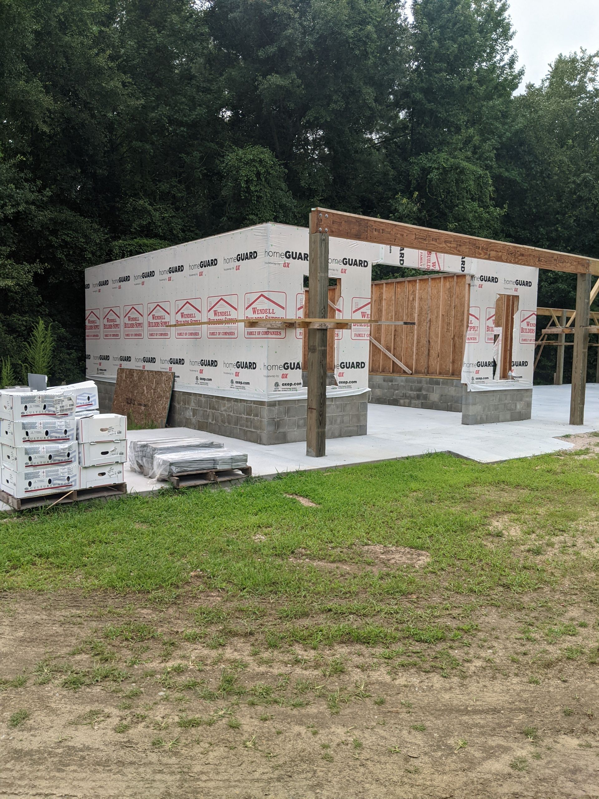 A building under construction with white wrap on its exterior, a concrete foundation, and wooden support beams.