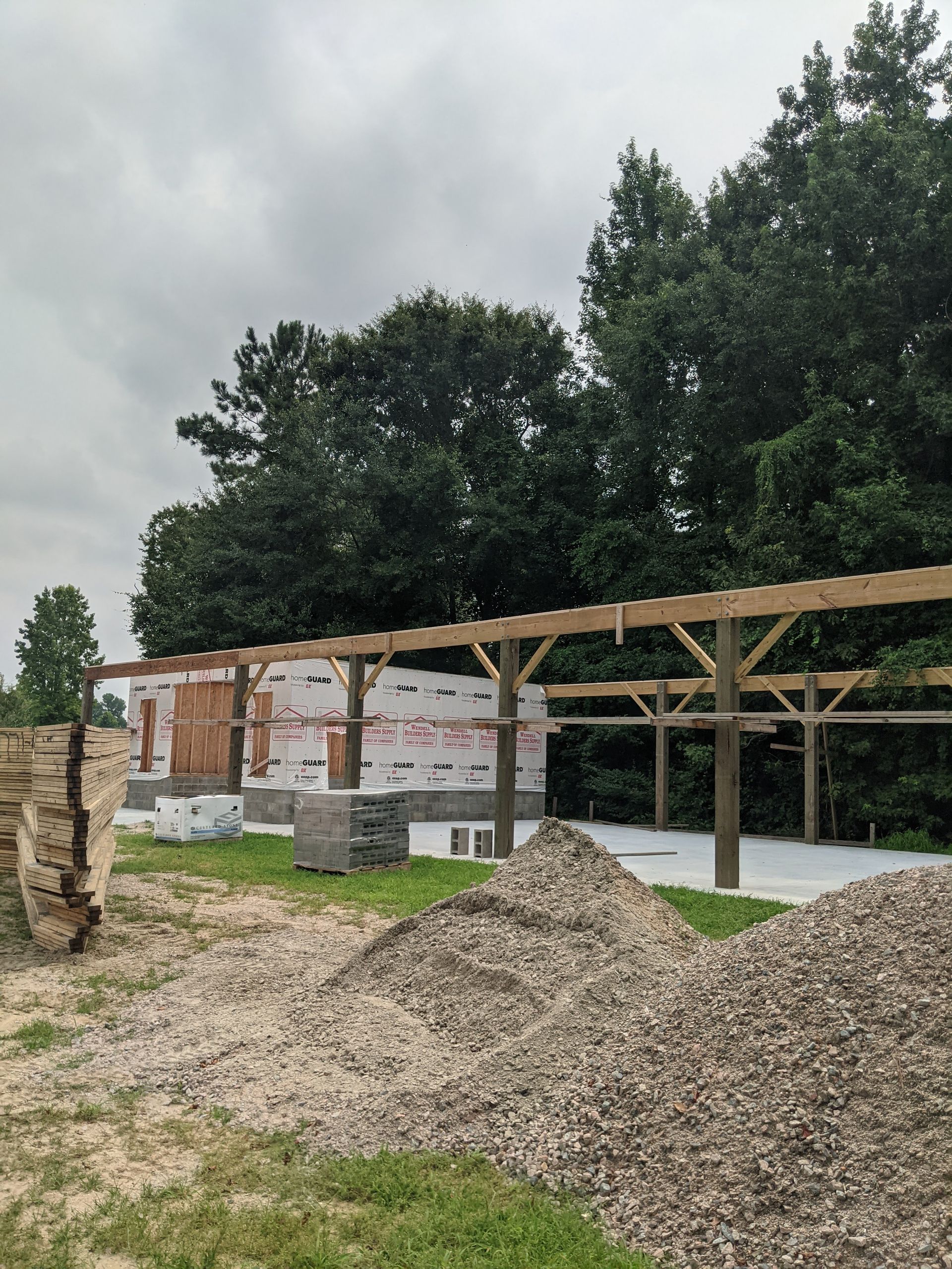 A building under construction with a wood frame pergola structure, piles of gravel, and trees in the background.