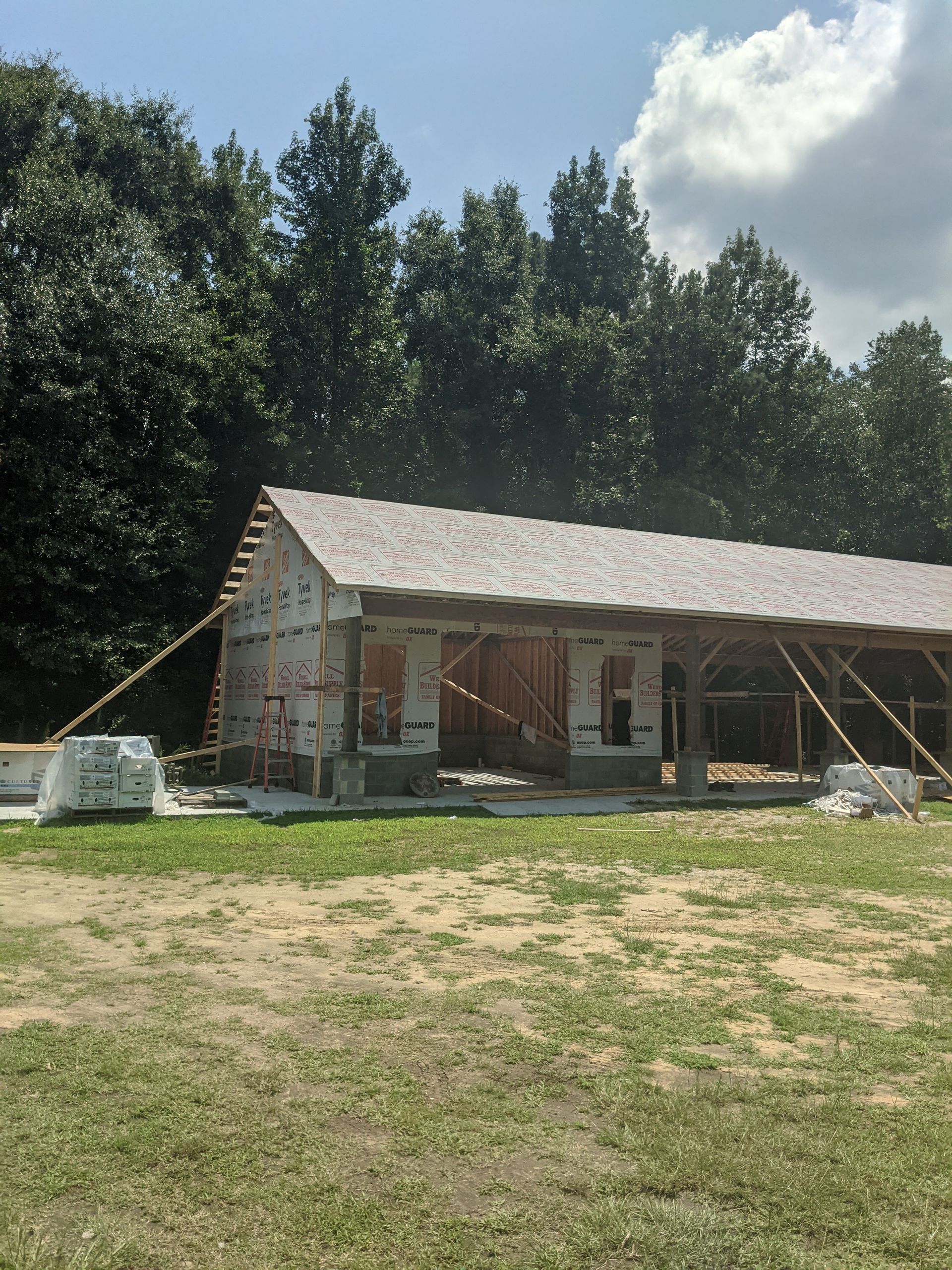 A partially constructed wood-frame building with a finished shingled roof, standing on a grassy lot near a line of trees.