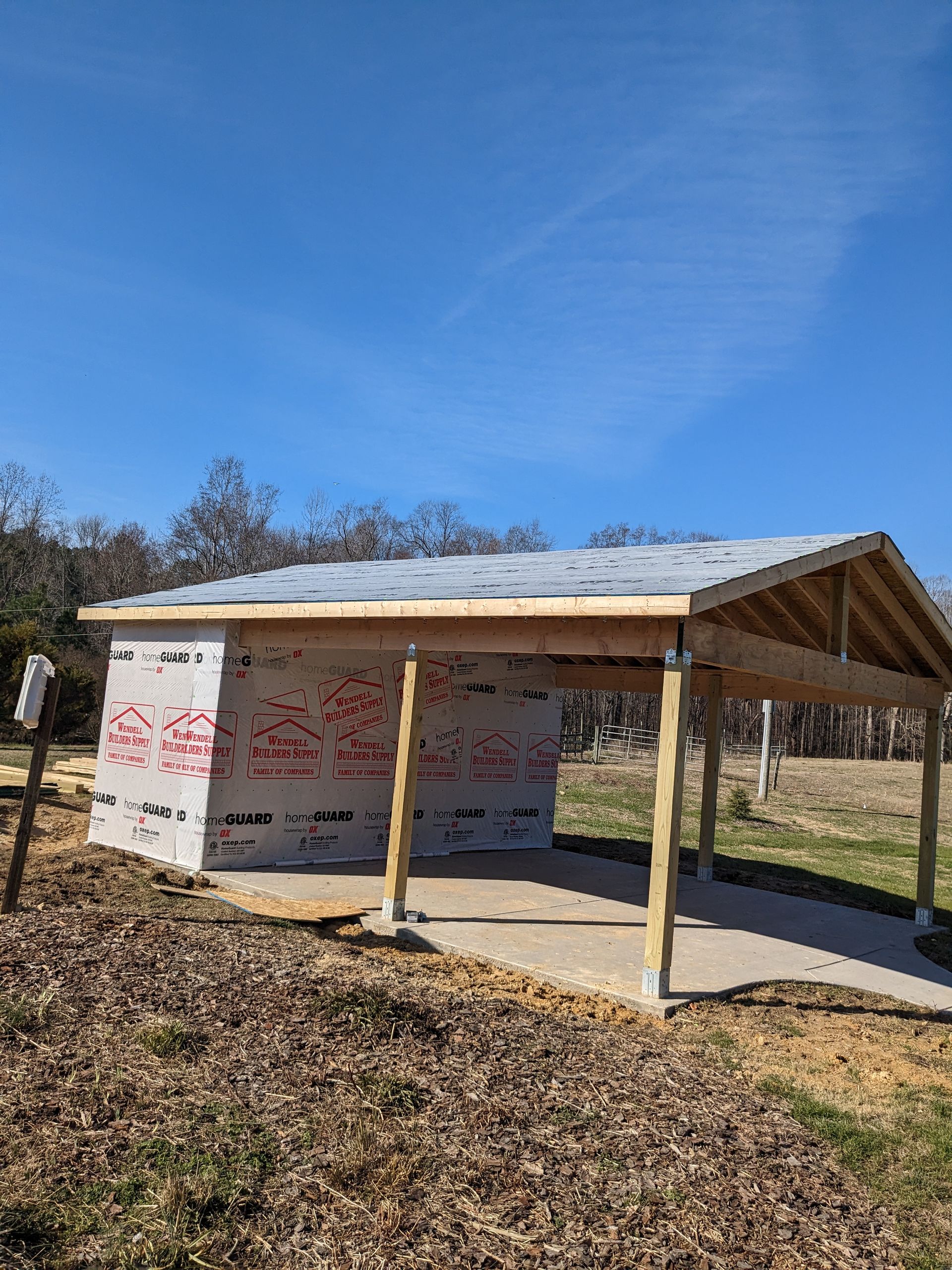 A partially constructed shed with a concrete floor, wooden frame, and white weather-resistant siding on a sunny, grassy lot.