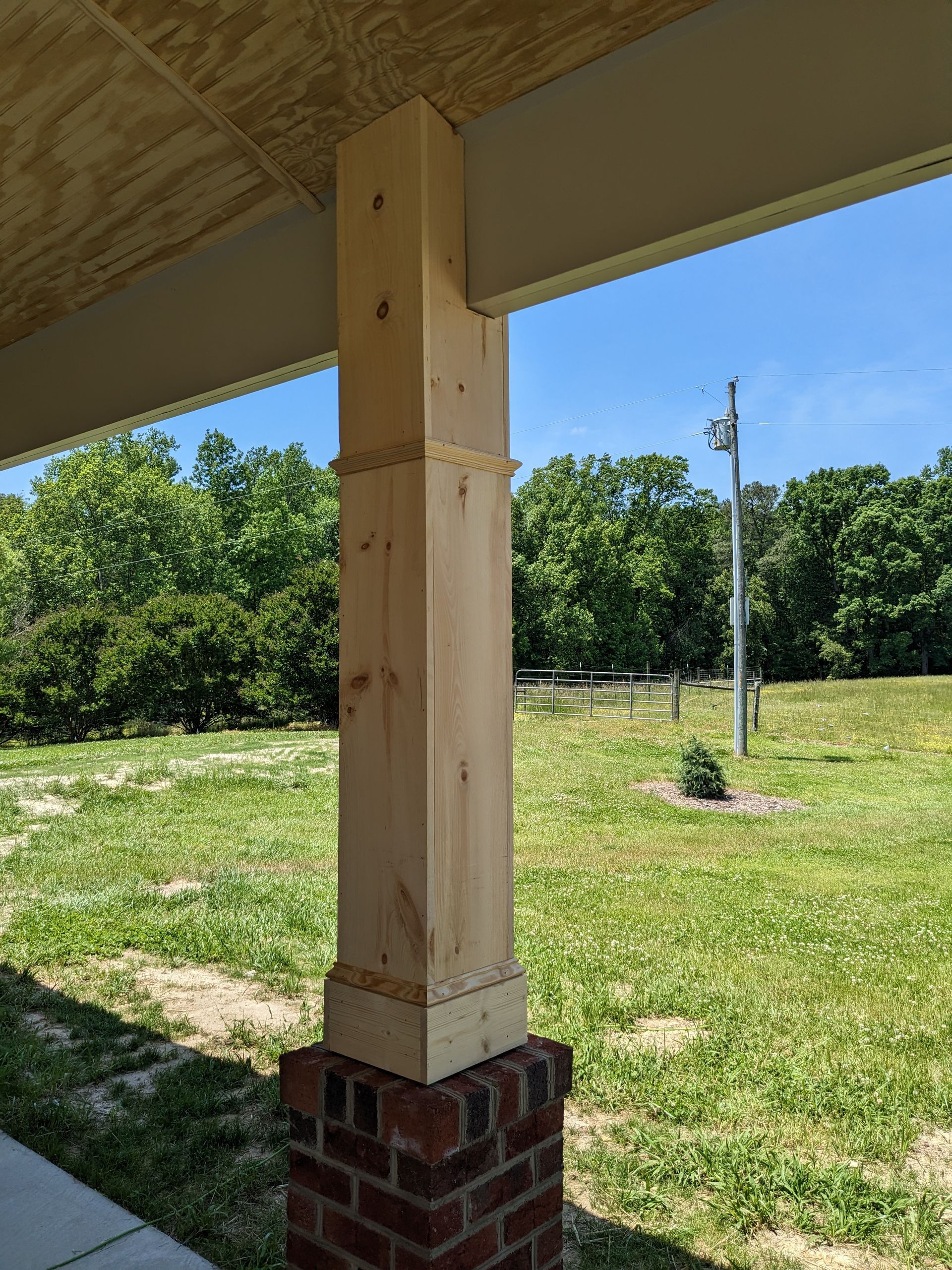 A wooden porch column resting on a brick base under a roof overhang, with a green, sunny landscape in the background.