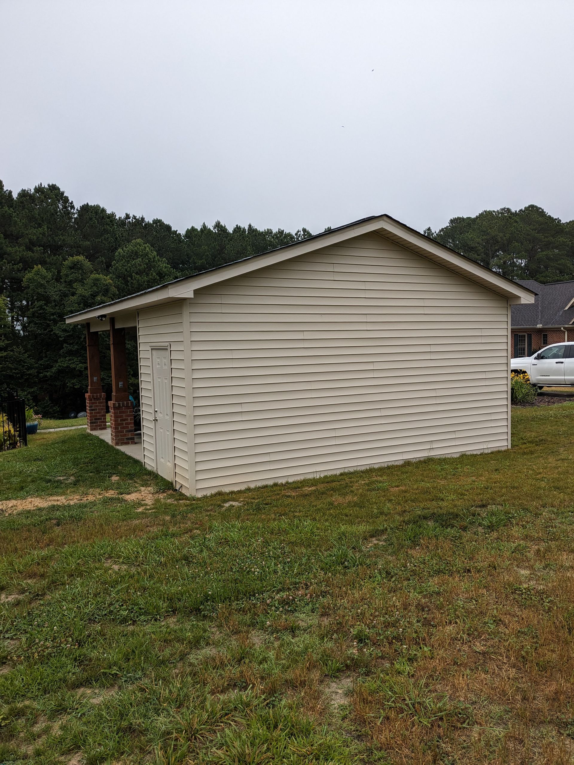 A small, light-colored shed with wavy, distorted exterior siding sits on a grassy hill near trees and a parked vehicle.
