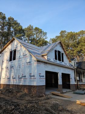 Partially built white house wrapped in Tyvek with exposed roof framing and brick foundation under a blue sky