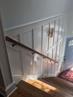 Sunlit staircase with white paneling, wooden handrail, and patterned rug at the bottom