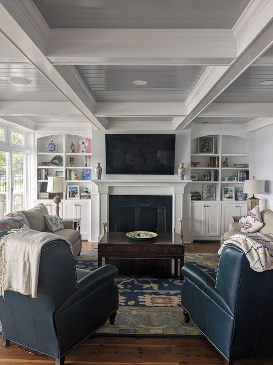Bright living room with white coffered ceiling, fireplace, TV, built-in shelves, and two blue armchairs facing it