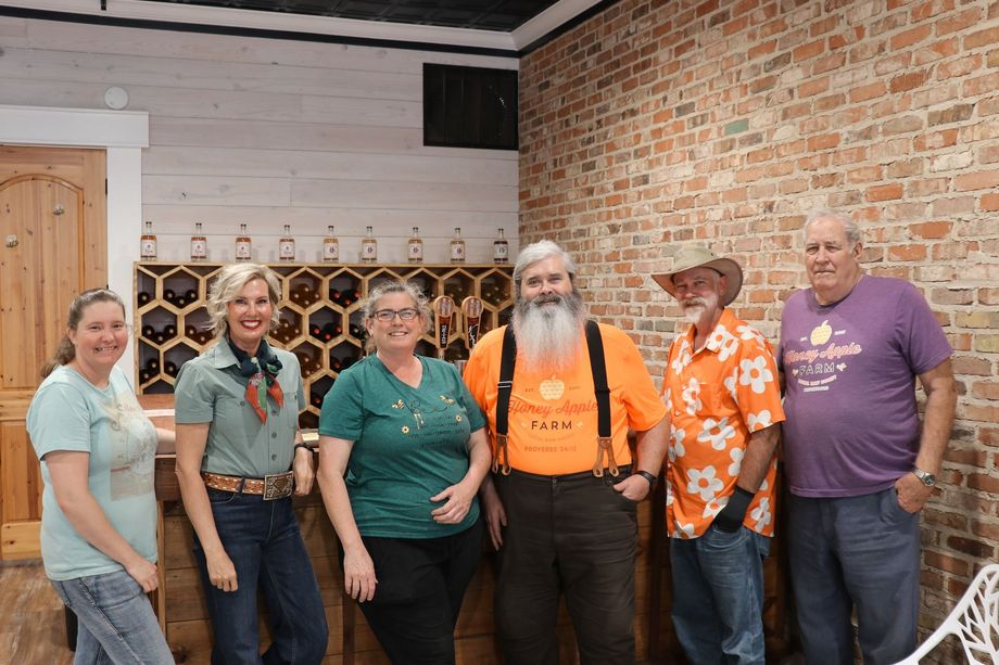 Six people standing together indoors in front of a brick wall, smiling at the camera.
