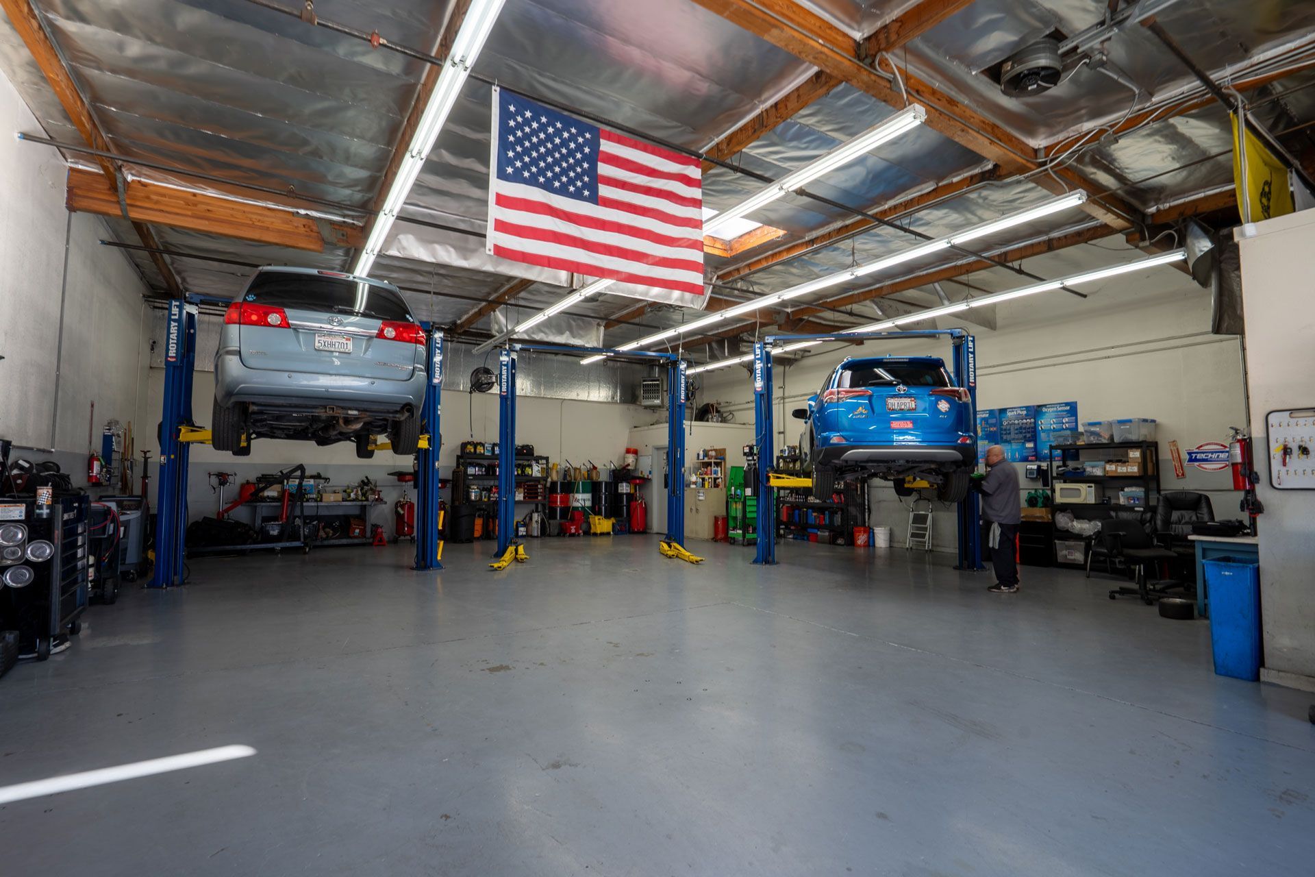 A car repair shop with two vehicles raised on lifts, American flag, tools, and a person.