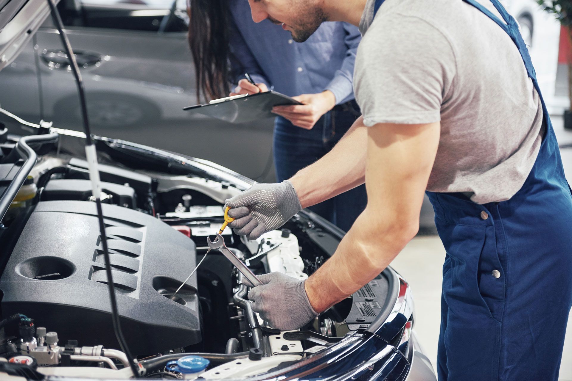 Mechanic in blue overalls working on car engine with a customer holding a clipboard in a garage.