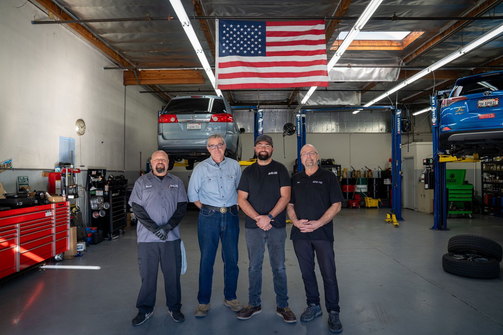 Four men pose in a car repair shop with a car on a lift and an American flag.