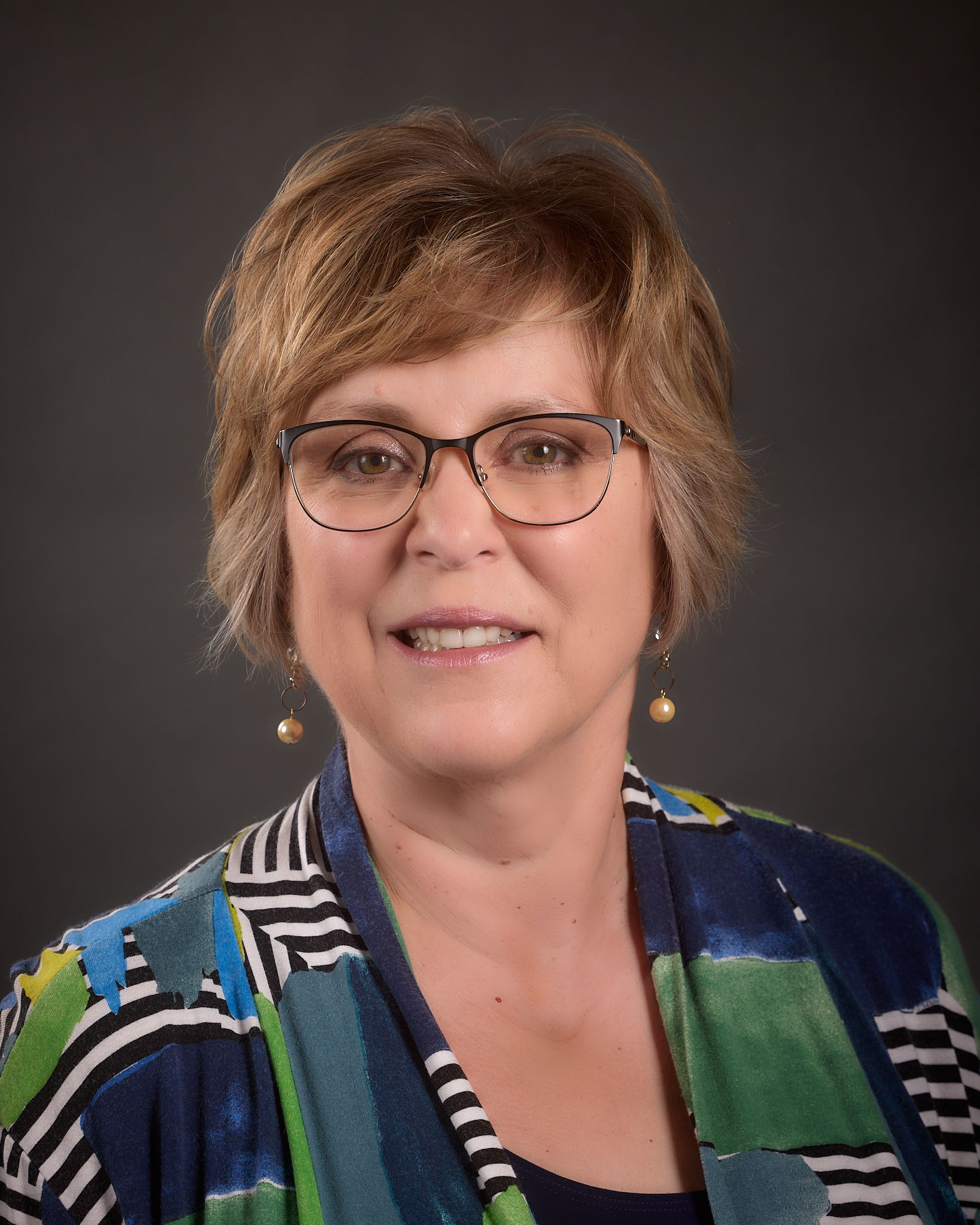 A woman wearing glasses and a colorful jacket is smiling for the camera.