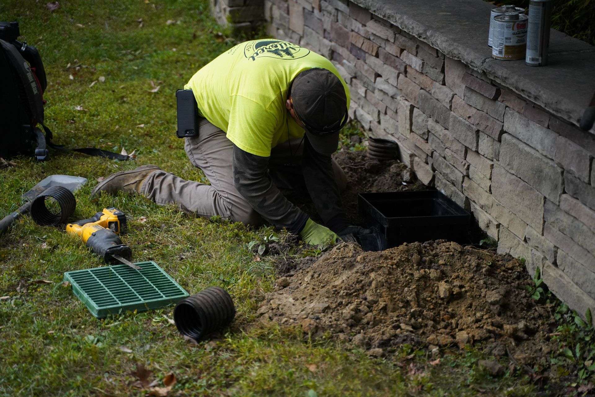 Worker in a neon jacket digging a hole beside a brick wall, with soil piles and tools on the grass.