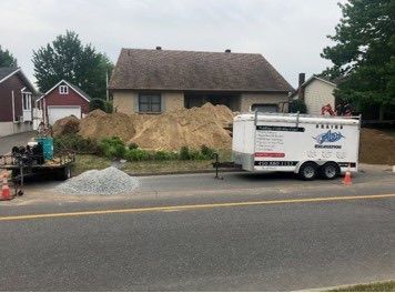 Chantier de construction avec des tas de sable, du gravier et une remorque blanche au bord d'une route devant une maison