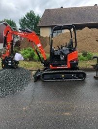 Une mini-pelle rouge creuse du gravier à côté d'une maison sur une allée mouillée.