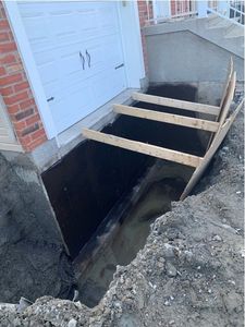 Puits de lumière ouvert au sous-sol, jouxtant une maison, avec des planches de bois enjambant une tranchée.