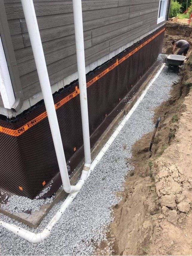 Fondations de maison recouvertes d'une membrane d'étanchéité à côté d'une tranchée de gravier et de descentes de gouttière blanches.