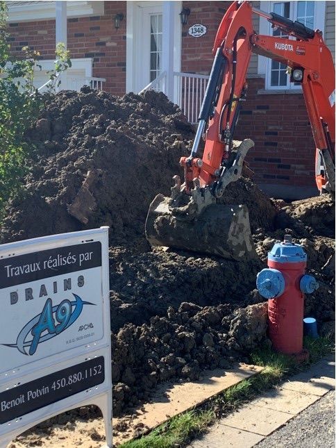 Une pelleteuse orange creuse un grand tas de terre à côté d'une maison en briques et d'une borne d'incendie.