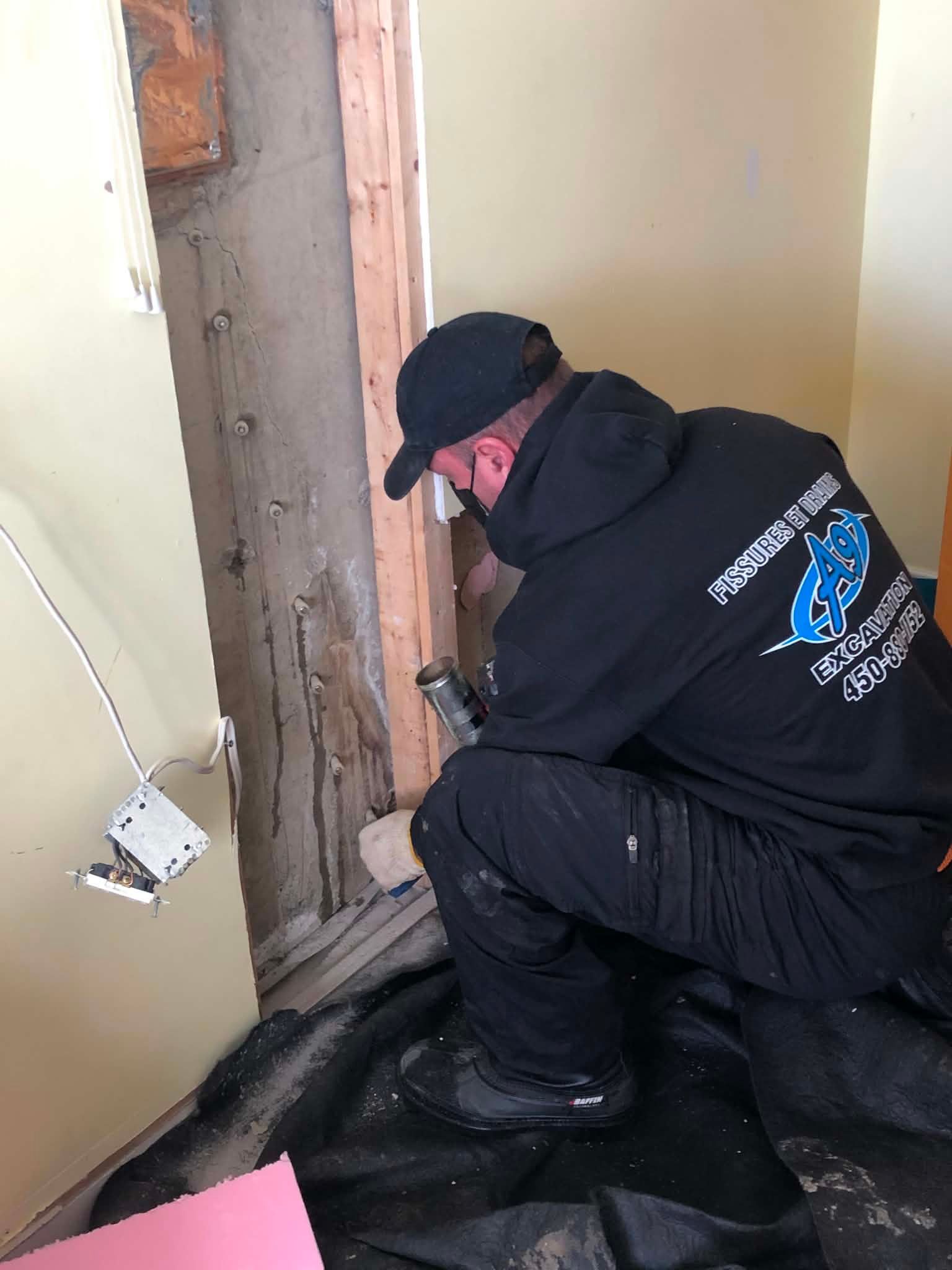 Un ouvrier en uniforme noir est accroupi près d'une porte, réparant un mur endommagé dans une petite pièce.
