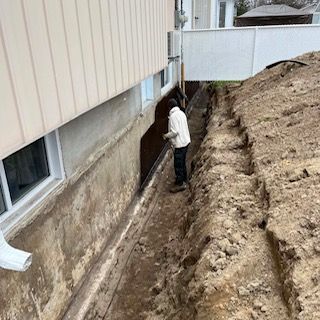 Un ouvrier, dans une tranchée à côté des fondations d'une maison, inspecte un mur excavé et le sol.