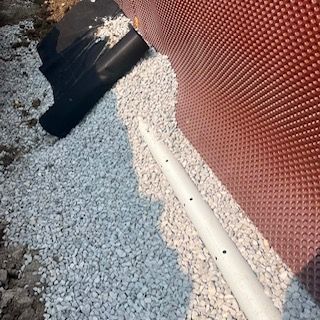 Gros plan sur du gravier à côté d'un mur rouge texturé, avec un tuyau blanc et une ouverture sombre dans le coin.