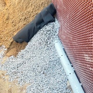 Matériaux de construction à côté des fondations : gravier, bâche noire, terre orange et tuyau blanc.