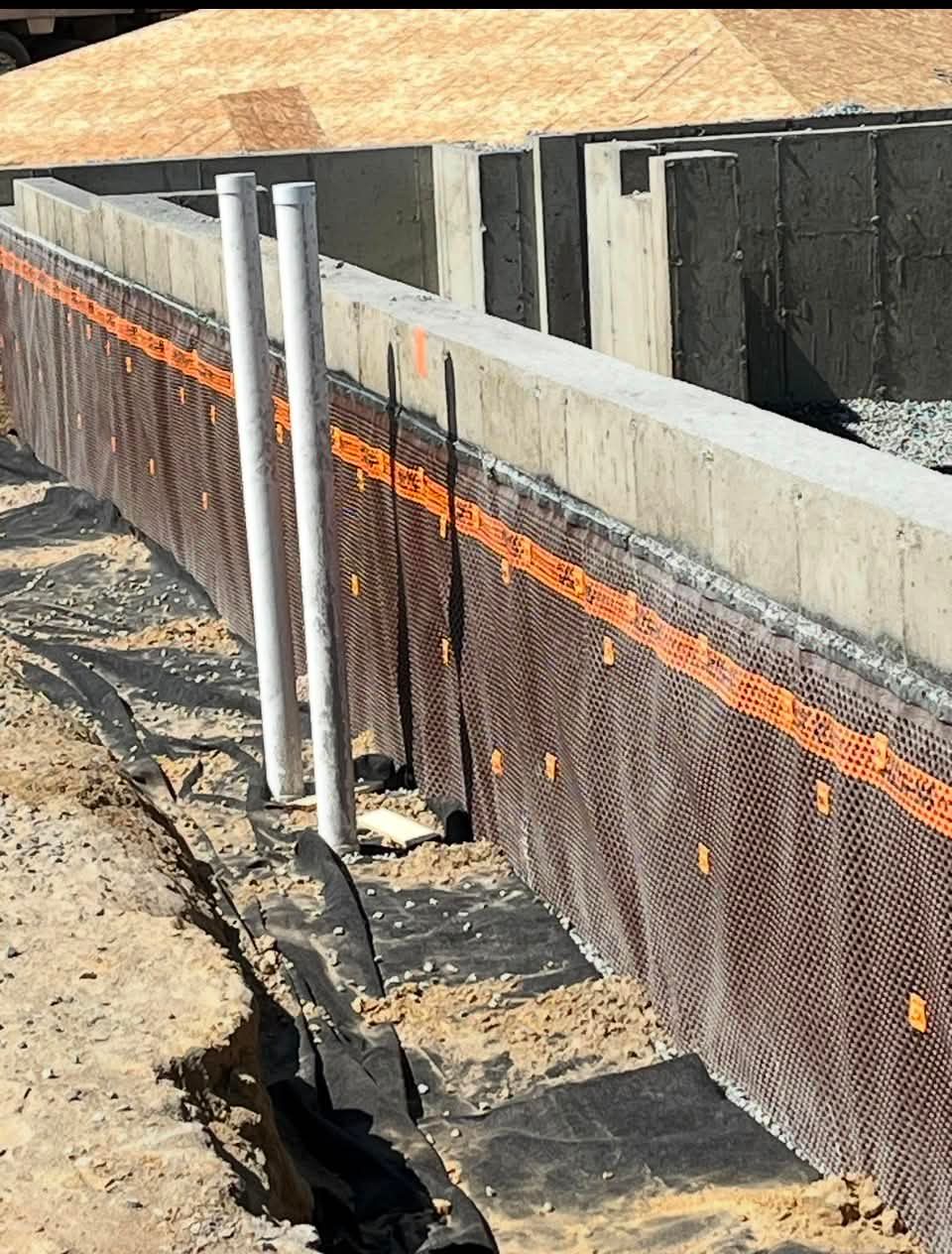 Mur de soutènement de chantier avec barrière en treillis orange et deux tuyaux métalliques à côté d'une fondation en béton