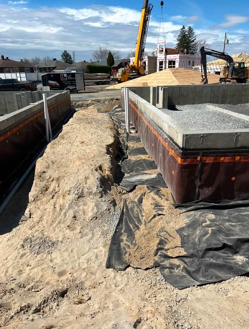 Chantier de construction avec excavatrice, tranchée de sable et murs de fondation en béton sous un ciel bleu