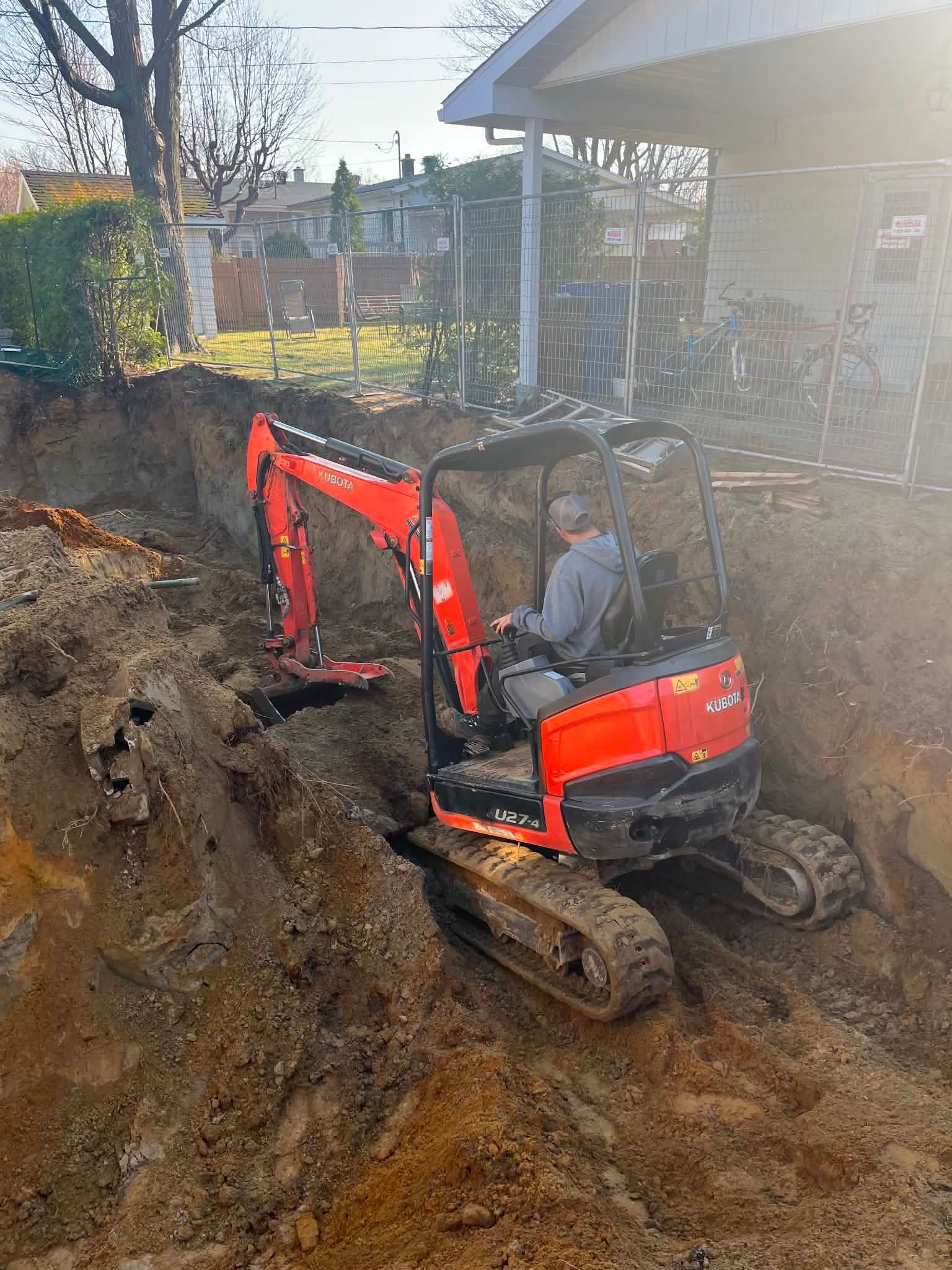 Une mini-pelle rouge creuse une tranchée de terre à côté d'une maison dans une cour