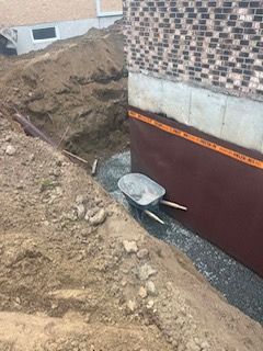 Fouilles de fondation le long d'un mur de briques avec du gravier, un monticule de terre et une brouette près du bord.
