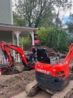 Une mini-pelle Kubota rouge travaille dans une cour boueuse près d'une maison, avec une autre pelle et un ouvrier à l'arrière.