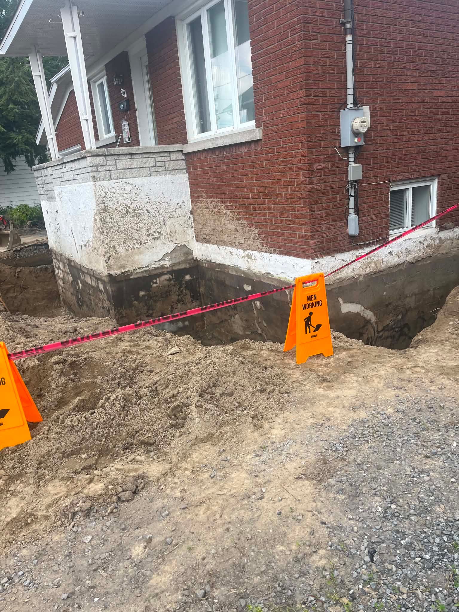 Travaux de terrassement à côté d'une maison avec fondations apparentes, cônes orange et ruban de signalisation rouge.