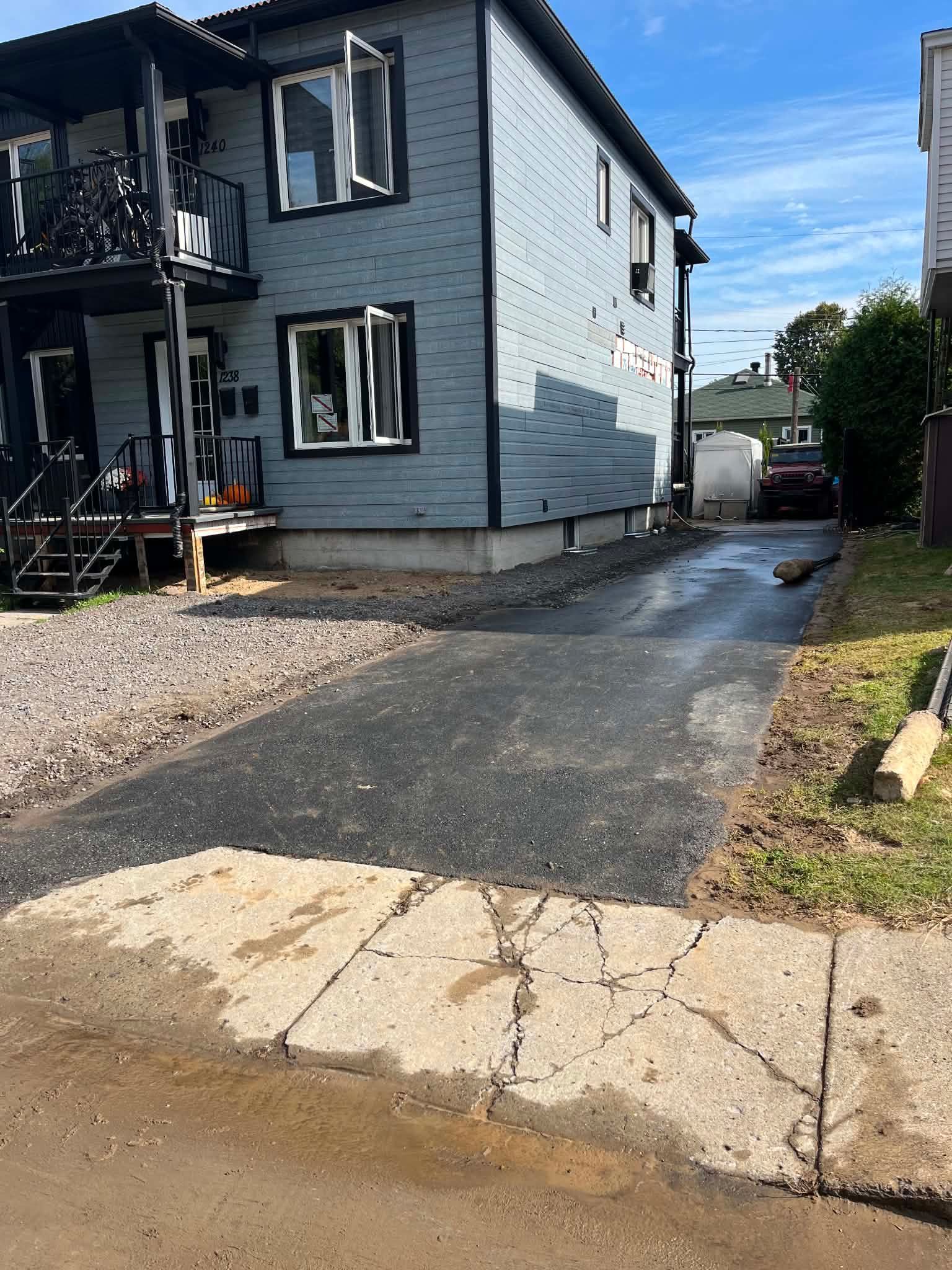 Allée étroite en asphalte longeant une maison bleue, bordée de gravier et donnant sur une rue mouillée.