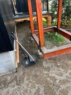 Terrasse extérieure avec une pelle appuyée contre un mur noir, des fondations en béton et des poteaux en bois apparents.