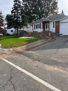 Allée résidentielle en construction à côté d'une maison blanche, avec du gravier et du matériel près du trottoir.