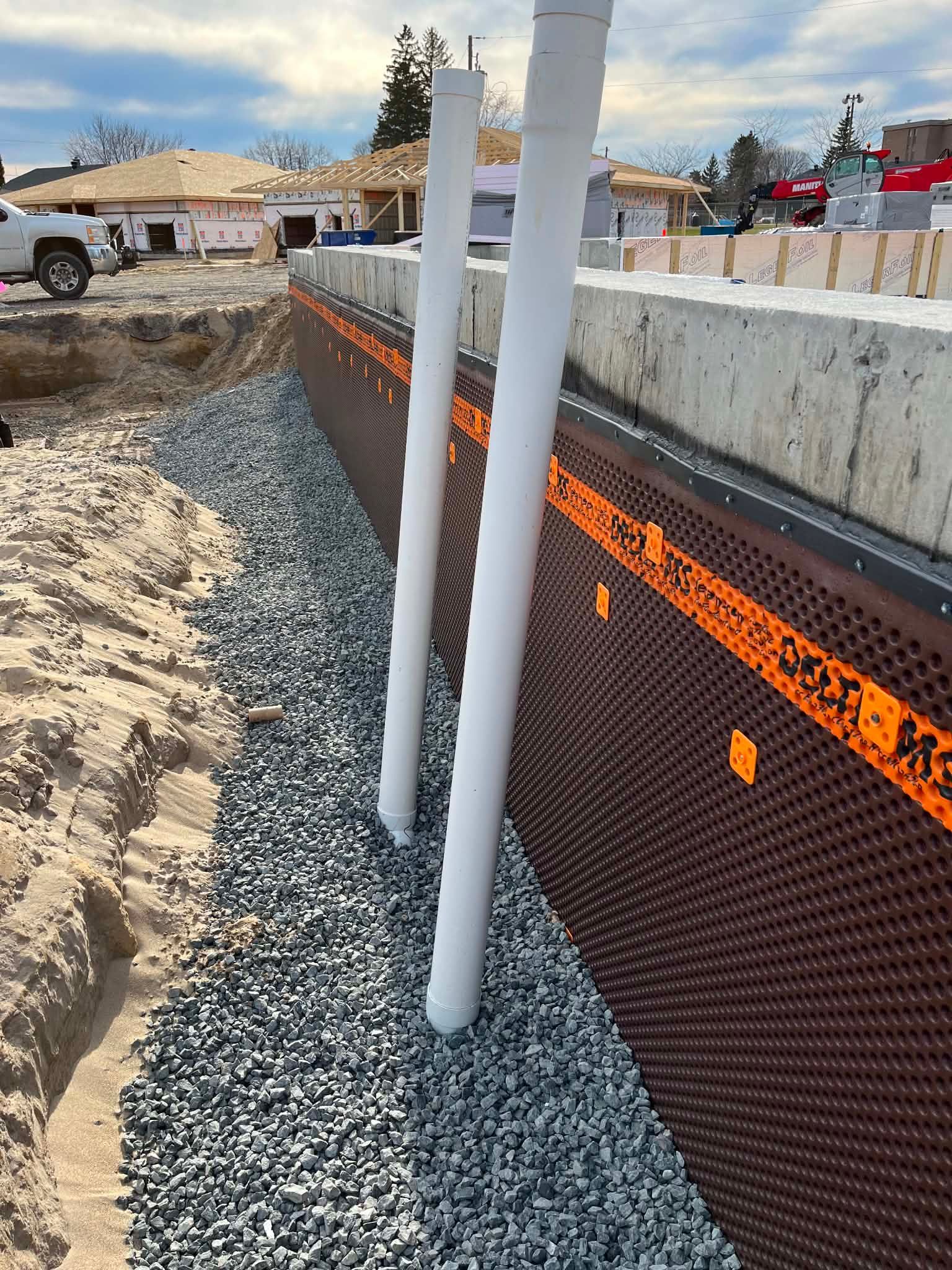 Tranchée de construction avec gravier, tuyaux blancs et clôture de sécurité orange le long d'un mur en béton.