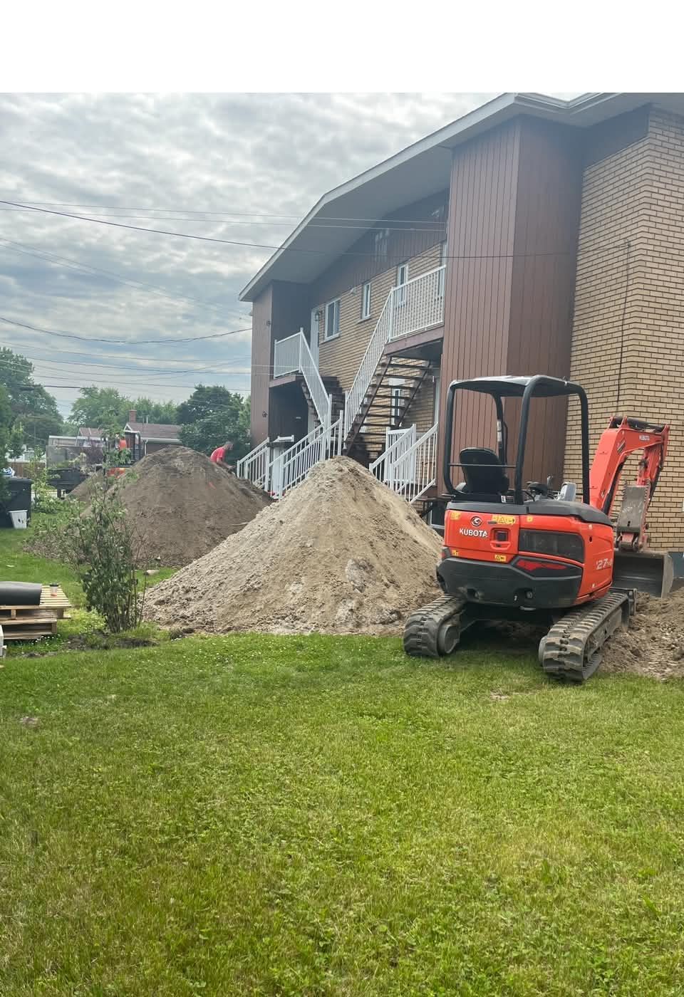 Une mini-pelle orange se trouve à côté d'un bâtiment partiellement effondré et d'un tas de gravier sur un terrain herbeux.