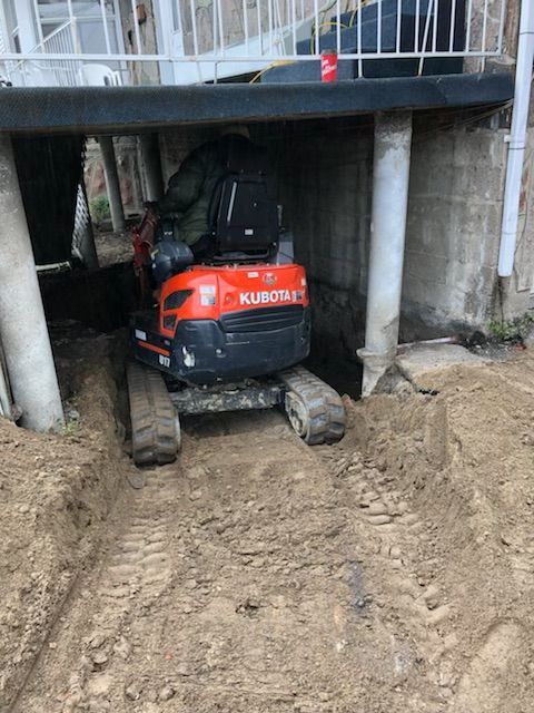 Une mini-pelle Kubota rouge creuse sous une maison, dans un sol boueux entre des piliers en béton.