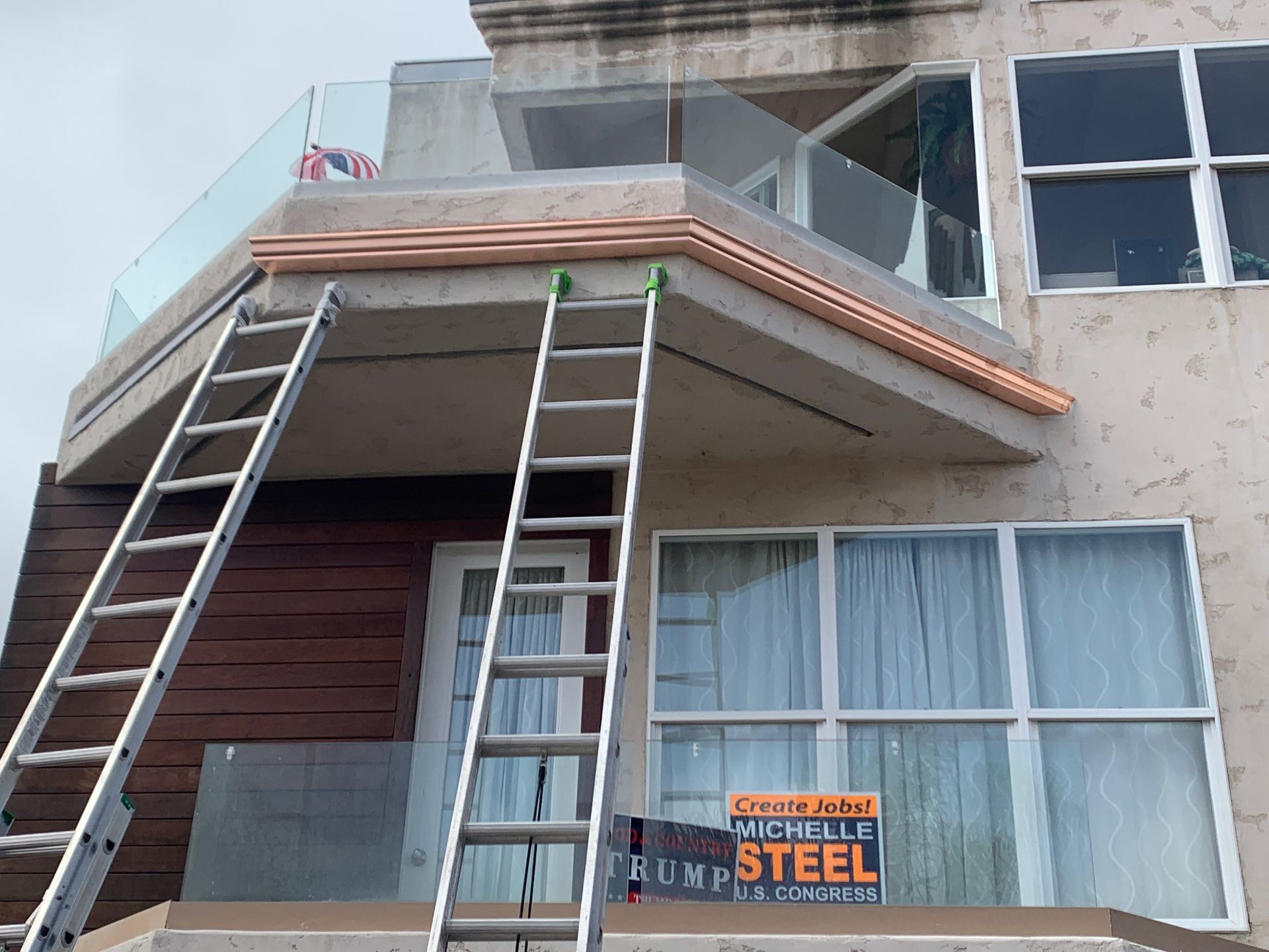 Two ladders against a building's balcony with glass railings, copper trim, and work sign at the bottom.