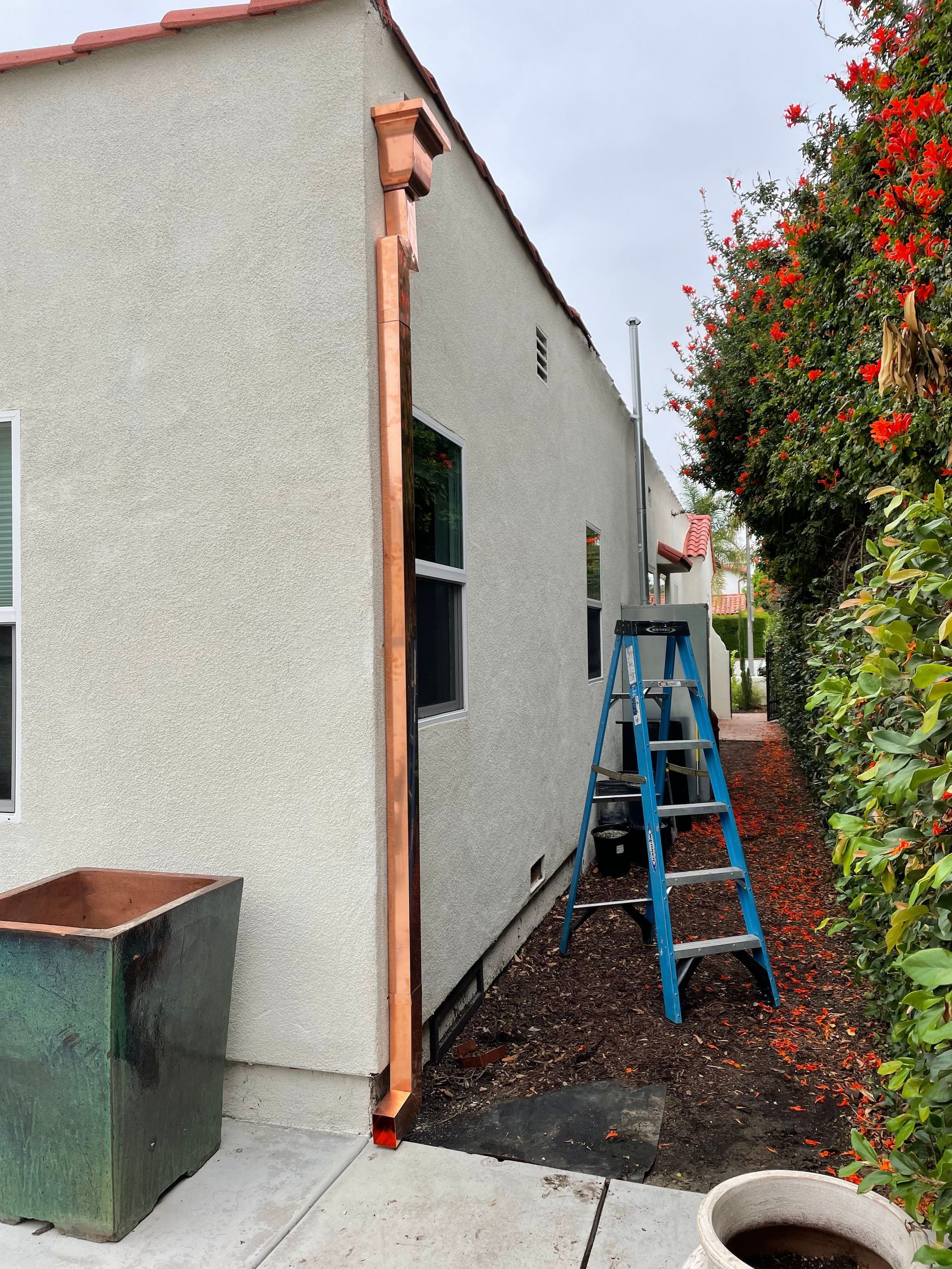 Copper rain gutter on a stucco building. A ladder stands nearby. A green hedge runs alongside.