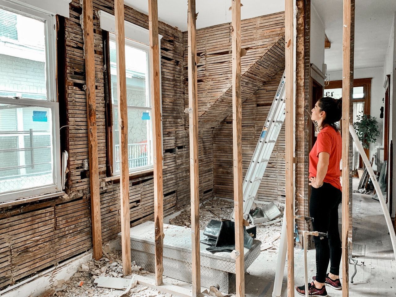 Interior renovation with exposed studs and brick walls; a person in an orange shirt stands beside a ladder.