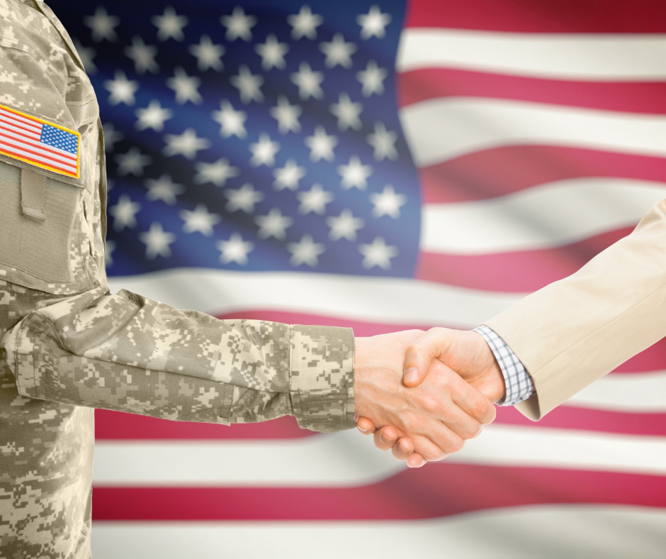 A soldier and a businessman are shaking hands in front of an american flag.