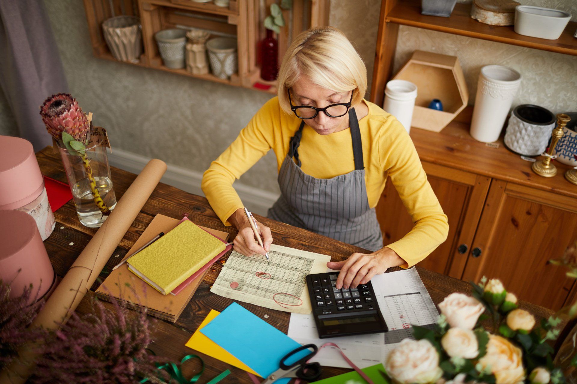 A woman is sitting at a table using a calculator and writing on a piece of paper.