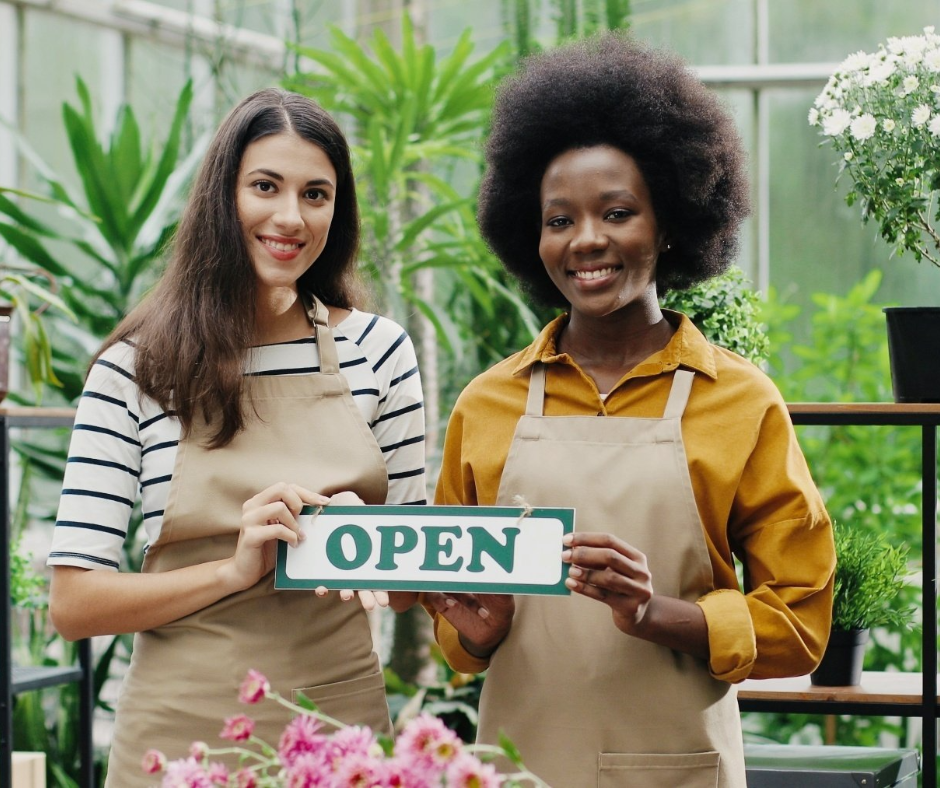 Two women are holding an open sign in a greenhouse.