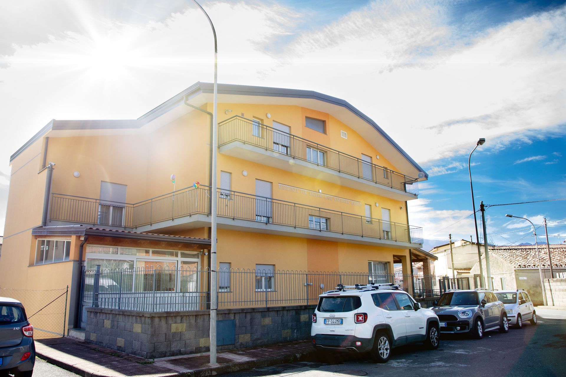 Edificio giallo con balconi, auto parcheggiate sulla strada, giornata di sole, cielo limpido.