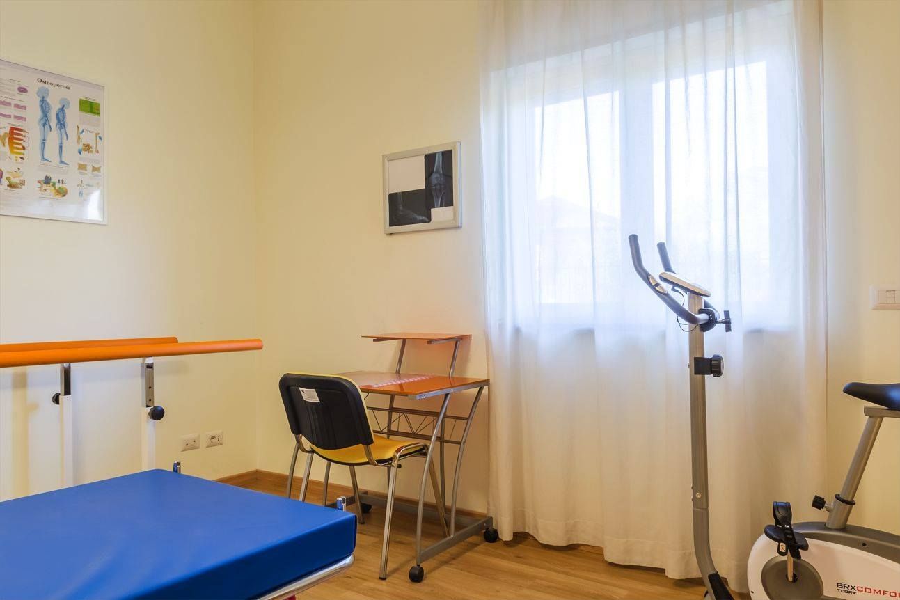 Sala di riabilitazione: tavolo regolabile, scrivania, cyclette, tabella spinale e finestra con tende.