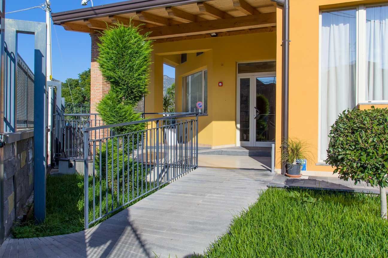 Edificio giallo con rampa e corrimano per l'accessibilità. Prato verde e un piccolo albero.