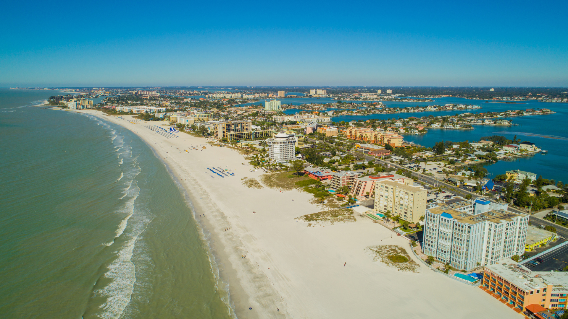 Aerial view of a wide sandy beach with turquoise water, shoreline buildings, and a coastal town in the distance