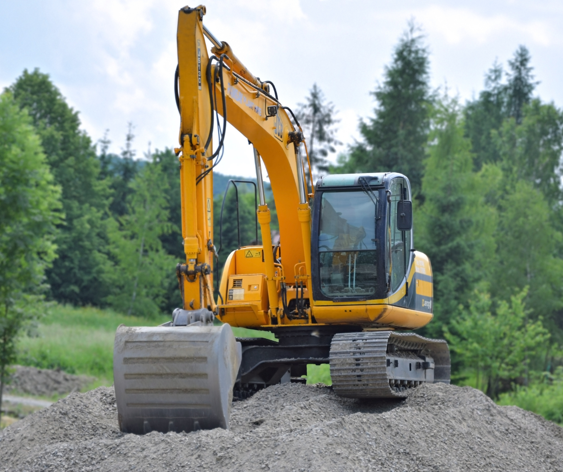 Yellow excavator on gravel, arm extended, against a backdrop of trees and a cloudy sky.