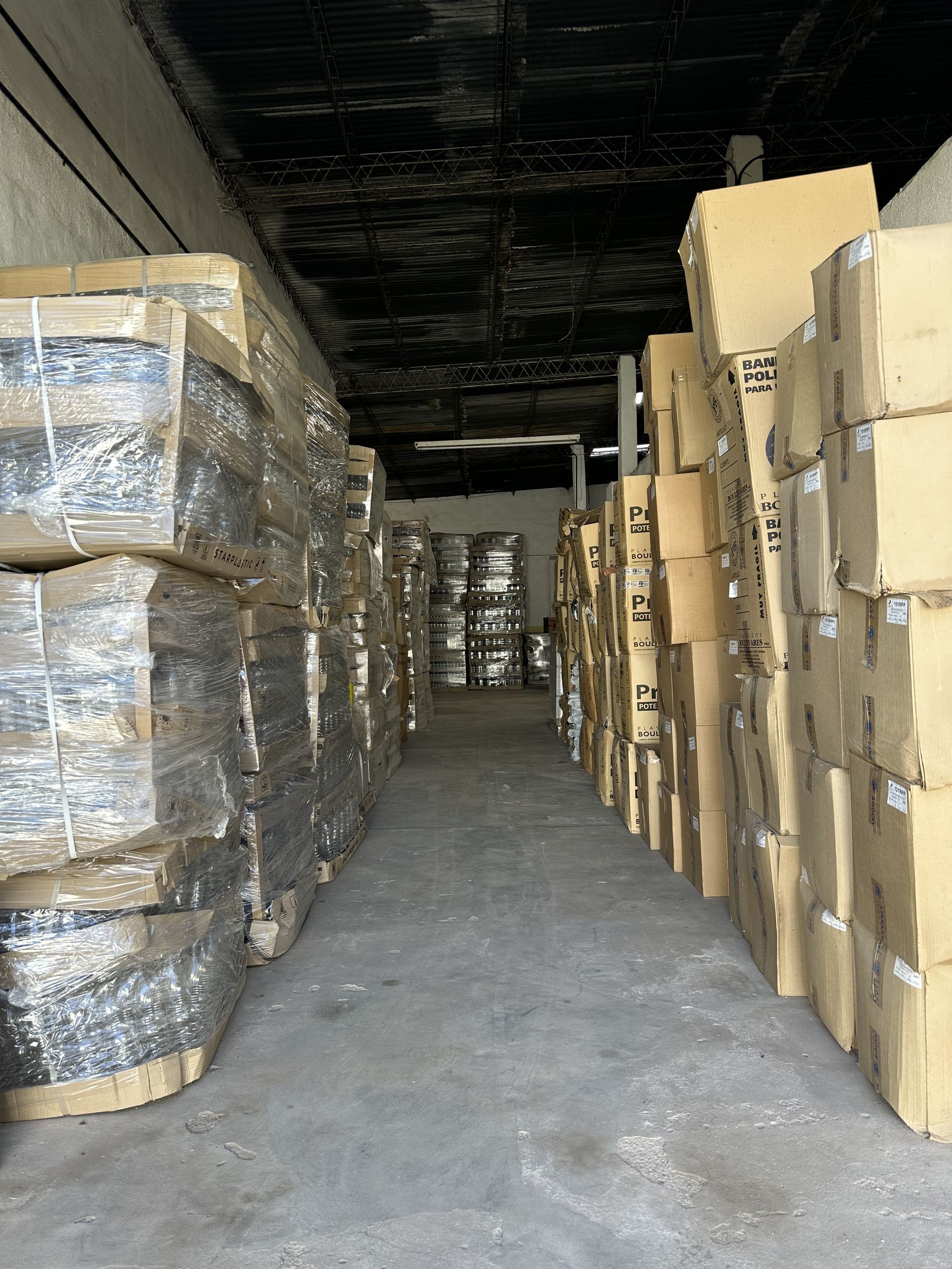 Pasillo estrecho de almacén con cajas de cartón apiladas a ambos lados y palés envueltos a la izquierda.